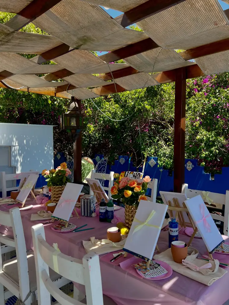 Sous une pergola de bois, une longue table nappée de rose alignant des toiles blanches sur chevalets, des pinceaux, des pots de peinture, des tasses en terre cuite et un bouquet de roses&nbsp;; au fond, le parapet bleu et un bougainvillier.