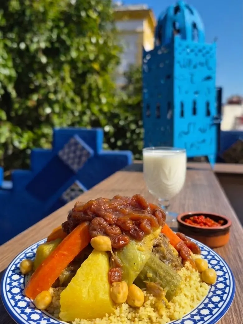 Sur une table de la terrasse, une assiette de couscous à motifs bleus garnie de carottes, courgettes, navets, pois chiches, viande et raisins secs confits&nbsp;; à côté, un verre de lben et une coupelle de harissa. Au fond, un décor de minaret peint en bleu vif.