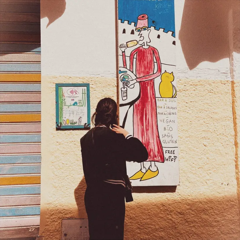 Sur un mur de la ruelle, un panneau peint à la main&nbsp;: un personnage en djellaba rouge coiffé d'un tarbouche tient un jus et une assiette, à côté d'un chat jaune et d'une liste manuscrite. Une passante, de dos, le regarde.