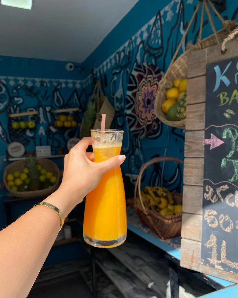 Une salle aux murs bleu pétrole entièrement couverts de dessins au trait noir&nbsp;; des paniers de citrons, d'ananas et de bananes sur des étagères, une ardoise à la craie, et une main qui tient une carafe de jus de mangue.