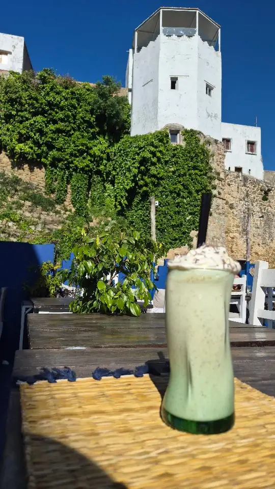 Depuis une table de la terrasse, la muraille de pierre couverte de lierre et, au-dessus, une tour blanche de la Kasbah sur un ciel bleu&nbsp;; au premier plan, un grand verre de milkshake.