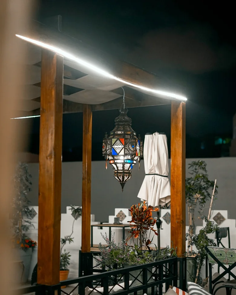 La terrasse de nuit&nbsp;: une pergola de bois éclairée par un néon, une lanterne marocaine en fer et verre coloré suspendue au centre, un parasol replié, des plantes et des banquettes.