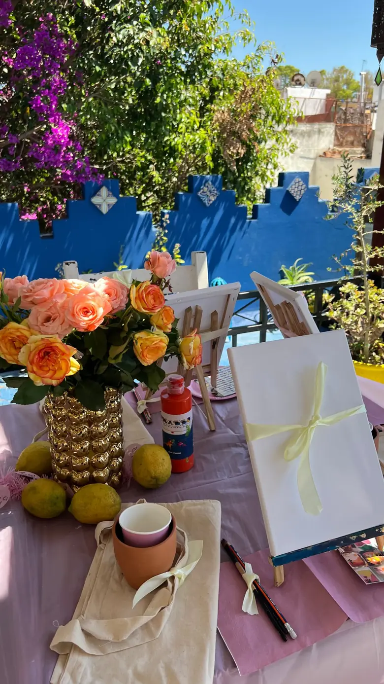 Une toile blanche montée sur un chevalet, posée sur une table dressée d'un tissu rose, avec des roses, des citrons et un pot de peinture&nbsp;; derrière, le parapet bleu de la terrasse et un bougainvillier.