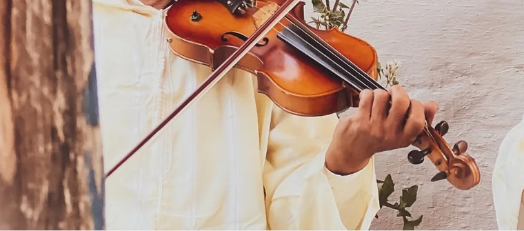 Cadrage serré sur un violon et l'archet&nbsp;; les mains d'un musicien en djellaba claire jouent devant un mur blanc où grimpe une plante.